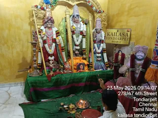 A photograph of a shrine room at KAILASA Tondiarpet with three deity murtis garlanded in white and orange flowers, standing before a green cloth and a gold-painted yellow wall. The left murti wears a red and gold turban and red garment, the central one a tall silver crown and green drape, the right one a silver crown and blue-green garment. A brass oil lamp, brass plates and small cups hold offerings in front, and a priest in white sits at lower right, lifting a small flame over a tray. At right stands a cutout poster of a smiling bearded man in a pink robe and red turban, wearing a red flower garland; a framed portrait of a woman sits nearby. A gold sign reads KAILASA TONDIARPET BHARAT.