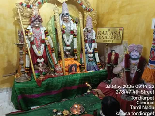 Photograph of a shrine room with yellow walls and a gold arch at KAILASA Tondiarpet. Three statues stand on a green draped altar: a central figure in saffron-orange with a tall crown and garlands, flanked by two figures in red and green attire, one with a turban and gold ceremonial necklaces. A small framed photograph sits in front of the central statue, and a gold sign reads KAILASA TONDIARPET BHARAT. To the right, a painted or printed life-size figure of a smiling man wearing a pink-red turban, red garlands and rudraksha-style beads sits against a red cloth, with a flower garland beside him. In the lower foreground, a person's hand holds a lit oil lamp over a brass tray with flowers, kumkum and brass vessels. The timestamp reads 23 May 2025 09:14:22, 278/47 4th Street, Tondiarpet, Chennai, Tamil Nadu. A partially visible head in the foreground is the only physical person.