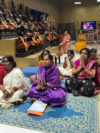 Women in saris sit on a blue and gold patterned mat in a temple hall, facing a row of black stone deity statues draped in red and gold cloth with garlands. A woman in a purple silk sari holds an orange folder on her lap, while a woman in a pink and black sari sits with palms pressed together in prayer. Other women in white, red, and orange saris sit nearby, and two more women in orange and yellow saris sit on chairs at the back. A wall-mounted television shows a screen with three figures, including a person in a crown and a turban. Plain white walls, folding chairs, and a brass lamp complete the setting.