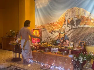 A priest in white dhoti with bare upper body stands at a puja altar, holding a lit lamp toward a deity picture framed in red with a golden crown image. Behind the altar a large backdrop shows a snow-covered mountain peak in warm sunset tones. A tall ornate figure in saffron-coloured turban, rudraksha mala and red-and-white garland is depicted on the backdrop beside framed photos. The altar holds fruit, flowers, brass bowls, coconuts and a placard reading KAILASA TAIWAN, with string lights and oil lamps below. Laghu puja ritual setting.