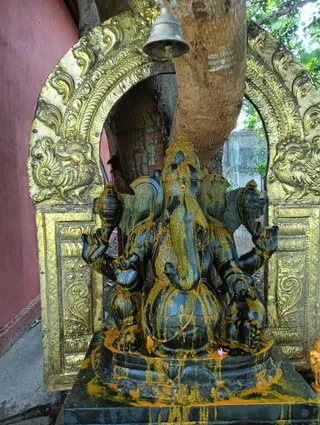 A photograph of a dark black stone Ganesha murti, a multi-armed elephant-headed deity, seated under a large tree trunk and a hanging brass bell. The image is smeared with yellow-orange turmeric or kumkum paste that runs down the statue and its base, and a small pink flower sits on the platform. The murti is framed by an arch of ornate embossed gold-coloured metal repoussé work against a red wall. A small golden figure stands at the right edge. No people are visible in the scene.