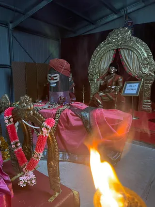 A photograph inside a plain metal-walled shed of a shrine. In the foreground, a dark polished Shiva lingam wears a red cloth draped over its top, a rich pink-and-gold draped cloth with a patterned border, a silver-and-red trident-like adornment, a pearl necklace and a gold-bordered base. Beside it, a pink and red garland of flowers hangs over a gilded ornate throne arch. Further right, a golden statue of a seated figure in red garments with a gilded ornate arch and pink floral garlands sits behind a small sign reading KAILASA AUSTRALIA. A flame from an oil lamp burns in the bottom foreground. No physical people are visible.