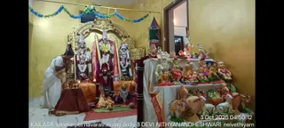 A worship scene in a yellow-walled shrine room with a white marble floor. A priest in a white dhoti and shawl stands at left with hands folded in prayer before a small offering table set with brass lamps and coconuts. Behind him, three ornately garlanded deity murtis stand under a golden arch, the central figure dressed in a red and gold sari-like cloth with a tall silver crown and a yellow garland. Strings of blue and green paper decorations hang above. On the right, a long display shelf holds rows of colourful painted clay dolls and figurines, including figures of animals and seated devotees. A lit oil lamp and a small kumkum-marked panel sit near the center, and a sign at the top right reads KAILASA THONDAIYARPET.