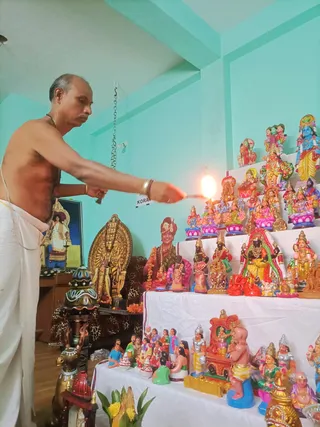 A photograph of a bare-chested man in a white dhoti performing a Navaratri golu display, holding a lit flame or lamp toward a tiered arrangement of painted clay dolls and deity figures on white-draped steps. A brightly coloured Hindu deity figure with blue skin stands at the top right, and a gilded multi-armed deity murti sits at left beside a framed picture. A seated figure in a red and gold turban, garlanded with flowers, is among the dolls at centre. Tiered shelves hold lotus-seated goddess figures in pink, green and yellow, and a small rath or chariot-shaped doll rests at lower right. A green shoot with yellow flowers sits in the foreground. The man is the only physical person visible.