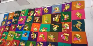 A photograph of a large grid of identical offering packets arranged on a pale wall. Each square of coloured cloth (orange, pink, green, purple, maroon and teal) holds a bundle of bananas tied with yellow cord, a leaf, pink flowers, a small heap of fruit or sweets, and a folded paper packet. No people are visible in the frame, and the arrangement suggests prepared offerings laid out for a puja.
