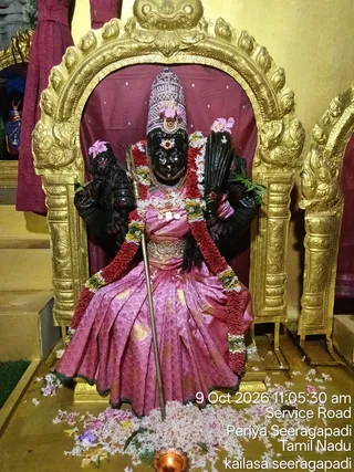 A black stone murti of a four-armed goddess seated on a golden throne with an arched gold frame and maroon backdrop. She wears a tall silver crown, a pink silk sari with gold border and waist belt, a red and white flower garland, and a trident held upright in one hand. Pink flowers adorn the upper arms, and white and pink petals are scattered on the floor before her, with a small oil lamp flame in the foreground. The photo is stamped with the date 9 Oct 2026, 11:05:30 am, Service Road, Periya Seeragapadi, Tamil Nadu, and kailasa seeragapadi.