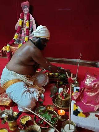 A bare-chested priest in a white dhoti with a gold-bordered waist cloth sits on a red floor covering, arranging a puja setup with brass vessels, a lit oil lamp, incense sticks, a bowl of mango leaves and red flowers, yellow and red powders, and a brass kalasha-style vessel with rose petals. He wears a white head cloth and rudraksha beads. Behind him, a red-turbaned framed portrait or cutout of a smiling figure in pink ornate clothing and a flower garland sits on a gold-and-red cushion against a red wall. A pink silk cloth and a rangoli-like pattern of coloured powders lie at right, and a wooden stick with red thread stands upright. This appears to be a devi puja ritual.