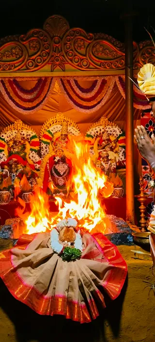 A large fire blazes in front of three decorated deity murti at an altar draped with orange fabric and garlands of marigold and tuberose. The central figure wears a red sari and gold crown, while the flanking figures are dressed in green and red with white flower garlands. A red and gold bordered cloth is spread on the ground in front of the fire, holding a coconut wrapped in white flowers and green leaves. On the right edge, a hand with a ring is raised in a gesture of offering beside a brass oil lamp, and a partly visible person holds a folded white cloth. The scene is lit by the fire against darkness and features ornate carved wooden arches overhead.