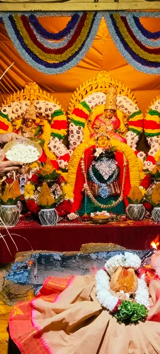 A photograph of a festival altar at KAILASA Hyderabad during a Navaratri devi ritual. Three goddess murti figures stand on a red-draped platform, adorned with yellow and white flower garlands, gold crowns and gold arches, and red and green garland drapes. The central goddess wears a red sari, a green lower cloth, a silver breastplate and a gold necklace. Brass and clay pots with mango leaves and marigold flowers flank the figures. In the foreground a fire pit burns beside a coconut wrapped in white jasmine garland and green leaves, and a hand at the left holds a plate of white puffed offerings. Striped cloth bunting hangs above in blue, yellow, red and white.