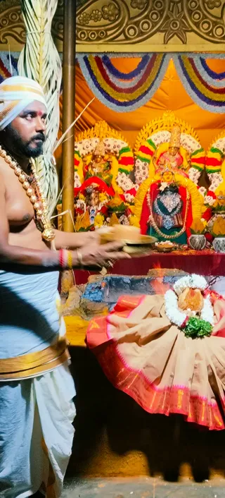 A priest in a white turban and white dhoti with a gold border stands bare-chested at left, wearing a gold beaded necklace and a red-and-gold bangle, holding a brass plate with offerings. Before a shrine draped with red, orange and multicoloured floral garlands, a deity murti in a silver-decorated form sits at the centre, with smaller murtis flanking it. In front, a brown-and-red sari-draped figure wears a white flower garland and green leaves beside a fire pit with a visible flame. Kalasha pots and flower arrangements sit on the altar table.