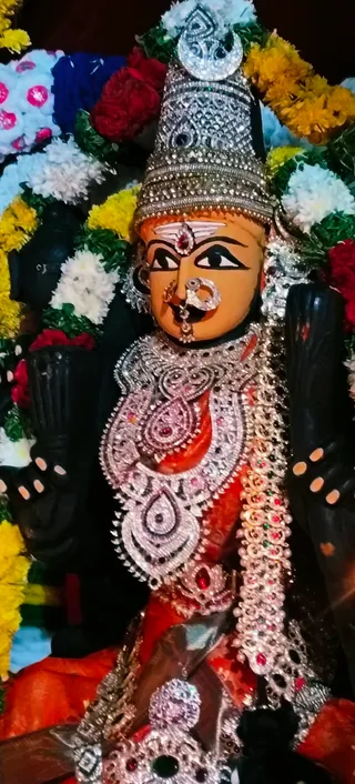 Close-up photograph of a dark-complexioned deity murti of a goddess. The face has painted red-orange skin, large black eyes, a red bindi and a vertical white tilak, and a large nose ring. Heavy silver and rhinestone necklaces and an ornate silver chest piece with red stones cover a red-orange garment. Garlands of white, yellow and red flowers frame the figure. No people are visible.