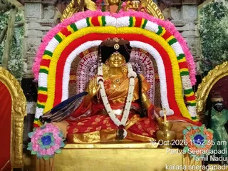 A seated golden deity murti, a goddess figure, is displayed inside an arched frame of garlands in yellow, red, white and green with a pink border. The murti wears a red and gold sari, a black crown-like headpiece, a white flower garland and gold necklaces, and raises one hand in blessing. Behind it are red and white patterned panels and a green painted tree backdrop. A brass kalasha or lamp stand and a pink and blue floral decoration sit at the lower corners. A small green-robed figure stands at the right edge, partly cropped. The timestamp reads 16 Oct 2026 11:52:35 am, with Periya Seeragapadi, Tamil Nadu and kailasa seeragapadi printed in the corner.
