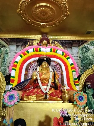 A photograph of a golden statue of a seated female deity inside a shrine, garlanded with a white jasmine garland and a multicoloured floral arch of pink, yellow, red, white and green flowers behind her. She wears a red and gold sari, a dark head covering with a gold crown-like ornament, and her right hand is raised in abhaya mudra beside a brass oil lamp. Above her sits a smaller golden figure in a maroon turban. Decorative fabric flowers in pink and orange-blue sit at the sides, and a dark-skinned figure in green clothing stands to the right. A timestamp reads 16 Oct 2026 11:52:13 am, with the location Periya Seeragapadi, Tamil Nadu. The statue is a deity murti, not a living person.