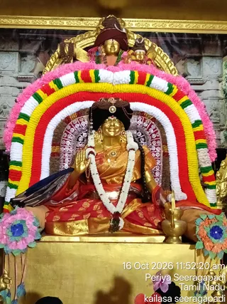 A seated golden deity murti is the focus of a temple shrine, dressed in a red and gold silk garment with a white garland hanging from its neck. Its right hand is raised in blessing and it holds a kamandalu-like vessel in its lap area. Behind it, an elaborate arch of concentric garlands in pink, white, yellow, red and green frames the figure, with a gold-framed backdrop showing temple architecture. Small golden figures appear above the arch and decorative artificial flowers in pink, blue and orange hang at the sides. A timestamp reads 16 Oct 2026 11:52:20 am with the location labels Periya Seeragapadi, Tamil Nadu and kailasa seeragapadi. No people are visible.