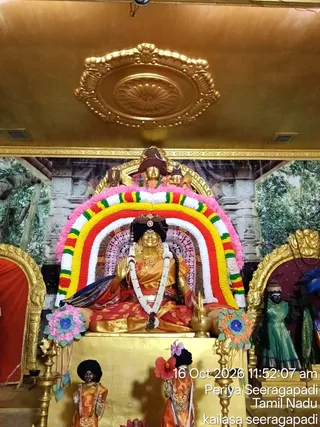 Photograph of a gilded deity murti seated in a shrine beneath a gold ceiling medallion. The seated golden figure wears a red and gold sari, a white flower garland, and a dark crown, with raised hand in abhaya gesture. Behind it is an arch of marigold and ribbon-like garlands in pink, white, yellow, red and green, framed against a painted backdrop of temple architecture and trees. Smaller golden figures stand at the top of the arch, and to the right a dark-faced figure in a green sari stands beside a blue floral decoration. Brass oil lamps stand on either side and decorative floral pieces sit at the base, with two small statues in orange garments below. No living people are visible.