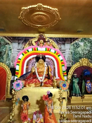 Interior shrine with a seated golden deity murti in a sitting posture, hands raised in abhaya-like gesture, wearing red and gold garments and a rudraksha-style garland, framed by a large arch of pink, white, yellow, red and green flower garlands. Flanking the central figure are smaller standing figures in orange robes, dark-skinned figures in a green sari and blue-trimmed attire, a gilded arch and a painted backdrop of trees and temple architecture. Brass oil lamps stand on either side of the golden pedestal, and a golden ornamental ceiling medallion hangs above. Two standing attendant figures with dark topknots hold a red flower and a staff near the base of the altar. This is a photograph of the shrine with no living people visible.