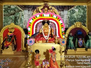 A decorated shrine with a seated deity murti dressed in a gold and red sari, its hands raised in blessing, framed by a large arch of red, yellow, white and pink flower garlands. Smaller figures in orange and saffron garments and tall black hats stand on the golden base before it, with brass oil lamps on either side. Flanking niches hold more figures: one in red with a flower garland, others in green and blue robes. Painted forest backdrop and a carved temple wall sit behind. Floor rangoli patterns fill the lower corners. No physical people are visible; the figures are all statues.