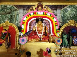 A photograph of a gold-coloured deity murti seated in a shrine decorated with a multicoloured floral arch of red, yellow, white and pink garlands. The central seated figure wears a red and gold garment, a white garland and a crown-like headdress, and holds one hand raised in a blessing gesture. Flanking niches hold a dark figure in a red and gold shrine on the left and a green-robed figure with a blue shield-like object on the right. Pink and orange floral decorations sit at the base, with a brass oil lamp beside the throne. The backdrop shows painted forest and temple imagery. A timestamp reads 16 Oct 2026 11:51:44 am, Periya Seeragapadi, Tamil Nadu, kailasa seeragapadi.