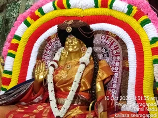 A golden deity murti seated in a decorated arch of white, yellow, red and green flower garlands at Periya Seeragapadi, Tamil Nadu. The murti wears a dark head covering with a golden ornament, orange and gold brocade clothing, a white garland draped over the shoulders, and a long dark braid. One hand is raised in a blessing gesture. A pink floral border frames the arch. The image carries a timestamp and place overlay.