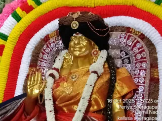 A gilded deity murti is seated in front of a multicoloured flower-garland backdrop of red, yellow, white and green. The figure wears a dark pleated headdress with a gold ornament, a red kumkum-style bindi, and large round earrings. Its dress is golden with a red and orange border, and it is decorated with white flower garlands and a gold chest pendant. Its right hand is raised in abhaya mudra. Behind it is a silver-embroidered panel with floral motifs and a rounded canopy. The photograph is stamped with the date and location 16 Oct 2026, 11:51:23 am, Periya Seeragapadi, Tamil Nadu, and kailasa seeragapadi.