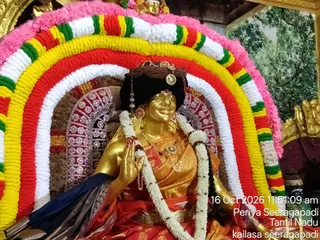 A gilded deity murti of a goddess is seated under a large arch of marigold and jasmine garlands in pink, yellow, red, green and white. She wears a dark crown-like headdress with a gold ornament, a gold necklace with pendants, and a gold-and-orange silk sari with a blue-and-gold border. Her right hand is raised in blessing, and a long white garland hangs across her body. Behind her is a red and gold patterned cloth and a painted backdrop of green foliage. A timestamp overlay reads 16 Oct 2026 11:51:09 am, Periya Seeragapadi, Tamil Nadu, kailasa seeragapadi. No physical people are visible.