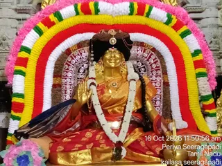 A golden deity murti seated in a padmasana-like pose inside a temple shrine, adorned with a red and gold silk garment, a white flower garland and gold jewellery. A bright arch of garlands in pink, yellow, red, green and white frames the figure, with a silver-embellished backdrop behind. The raised hand and seated posture suggest a blessing gesture. A visible timestamp and location overlay reads 16 Oct 2026, Periya Seeragapadi, Tamil Nadu, with a kailasa seeragapadi label. No people are present; the figure is a statue.