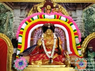 A golden deity murti seated in a shrine, decorated with a large floral arch of pink, yellow, red, white and green garlands. The figure wears a red and gold sari with a white flower garland draped over the shoulders and a dark crown-like headpiece. A brass oil lamp stands beside the murti, and a pink and blue flower arrangement sits at the lower left. Behind the arch, a golden framed backdrop shows a painted green forest scene and a small golden figure with a red turban above the arch. A timestamp reads 16 Oct 2026 11:50:52 am, Periya Seeragapadi, Tamil Nadu.