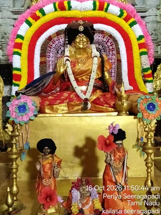 A golden seated murti of a female deity sits on a gilded pedestal under a multicolour floral arch of white, yellow, red and green garlands with pink petals. The figure wears a red and gold brocade sari, a white flower garland, a black headdress with a gold ornament, and gold jewellery. Her right hand is raised in blessing, and a brass kalasha sits to her right. In front stand two smaller orange-robed figures with black headdresses and red hibiscus flowers, and a small central murti garlanded with white and purple flowers. Brass lamps and paper flower decorations flank the scene.