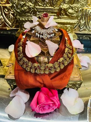 Close photo of a Shiva linga on a golden yoni pedestal adorned for worship. A rust-orange silk cloth drapes over the pedestal, with a gold multi-strand necklace of coin pendants and elephant motifs, and a white stone-and-pearl necklace with silver pearl drops above. Pink and white rose petals are scattered across the cloth and a metallic tray, with a single pink rose in the foreground. Behind is an ornate gold-embossed altar panel. No people are visible.