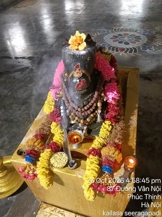 A photograph of a dark stone deity murti. It is decorated with pink and yellow marigold garlands, a yellow flower on its crown, layers of rudraksha malas, bells, and a red tilak-like mark on the forehead. In front stand a small brass oil lamp with a lit flame, a brass bowl of yellow grains, and a smoking incense stick. A second lit lamp sits at the right edge, and a brass vessel is visible at the far left. A colourful floral rangoli is on the floor behind. A timestamp overlay reads 23 Oct 2026 4:58:45 pm, with a location line reading 87 Đường Vân Nội, Phúc Thịnh, Hà Nội, and kailasa seeragapadi.