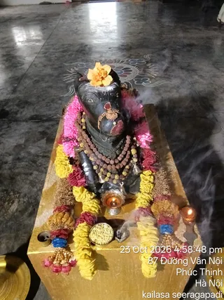A dark stone Nandi bull murti is the central subject, seated on a golden pedestal. It is decorated with a yellow and pink flower garland, a marigold-type orange flower on its head, a red-jewelled ornament on its forehead, and layered brown rudraksha malas with gold bead strands. Small brass oil lamps and a lit candle sit beside it, with a silver bowl of grains or powder and rising incense smoke. Brass bells and a brass plate are visible at the edges, and the floor is polished dark stone with a painted lotus-like motif. No people are visible.