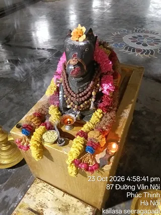 A photograph of a dark stone deity murti of Nandi, the bull, seated on a golden-brown pedestal inside a temple hall with a polished grey floor. The murti is adorned with pink and yellow flower garlands, a marigold on its head, rudraksha mala necklaces, a red tilak mark and a gold forehead ornament. A small brass oil lamp with a flame, a steel bowl of grains or rice, and a lit clay lamp sit beside it, with incense smoke rising. A brass vessel is at the left edge. No people are visible. A timestamp and location overlay reads 23 Oct 2026, 4:58:41 pm, 87 Duong Van Noi, Phuc Thinh, Ha Noi, and kailasa seeragapadi.