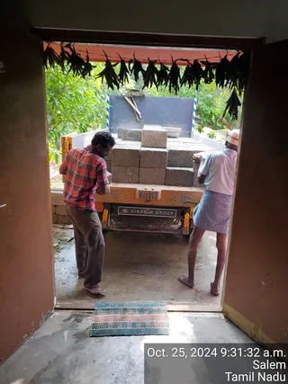 A photograph taken through a doorway of two men unloading concrete blocks from an orange truck parked in a yard. The man on the left wears a red and grey plaid short-sleeved shirt and brown trousers, and the man on the right wears a white shirt, a checked lungi and a white head cloth. Stacked grey hollow blocks sit on the truck bed, a dark blue-grey panel stands behind them, and a checked woven mat lies on the floor in front of the doorway. Green foliage and hanging dried leaves frame the top of the view. A timestamp in the corner reads Oct. 25, 2024 9:31:32 a.m. Salem Tamil Nadu.