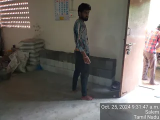 A photograph of an unfinished interior room with bare concrete floor and partly built walls of grey and white concrete blocks. A barefoot man with a short beard, wearing a striped long-sleeved shirt, dark trousers and a white bangle, stands near the middle looking toward the camera. Sacks are stacked against the left wall beside metal vessels, a wall calendar hangs above the blocks, and a second man in a red and white checked shirt and light trousers stands in the open doorway at right. A timestamp reads Oct. 25, 2024 9:31:47 a.m. Salem Tamil Nadu. No deity, abhishekam or ritual elements are visible.