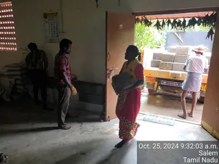 A photograph taken inside a plain, unfinished building with pale walls and a concrete floor, showing a construction-related activity. In the foreground, a woman in a red sari with a yellow-and-green patterned border carries a grey bundle toward an open doorway. To the left, a man in a red checked shirt, grey trousers and yellow gloves stands watching. Another figure in dark clothing stands near stacked sacks against the back wall. Outside the doorway, a man in a white shirt and checked lungi, with a straw hat, stands beside a yellow truck loaded with concrete blocks. Overhead hang dried leaves under a green canopy. A timestamp in the corner reads Oct. 25, 2024 9:32:03 a.m. Salem Tamil Nadu. Four people are visible.