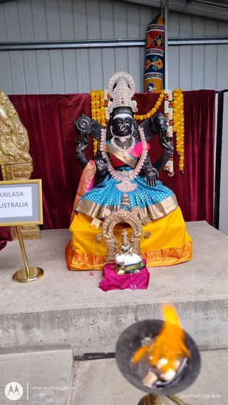 A photograph of a dark stone deity murti seated on a golden-yellow and blue silk-draped throne on a concrete platform, in front of maroon velvet drapes and marigold garlands. The murti has four arms, a jewelled crown with a halo, a red-and-white garland and gold ornaments, and holds objects including a discus-like emblem. A small golden shrine-like arch with a tiny figure sits before the throne on a magenta cloth. In the foreground, a metal lamp with a flame holds offerings. A gold-framed sign on a stand reads KAILASA AUSTRALIA. No people are visible.