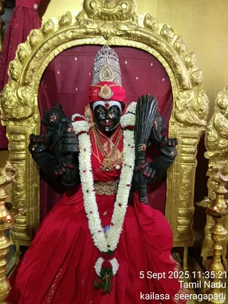 A photograph of a black-faced deity murti seated within a gilded arched shrine niche with a deep red cloth backdrop. The murti wears a red sari-like garment, a red headcloth beneath a silver crown, and layered gold necklaces and a gold waist belt. Multiple arms hold objects including a tall dark staff. A long garland of white flowers with red blossoms hangs from the shoulders to the lap. Brass oil lamps with lit flames stand on either side of the shrine. The photo carries a timestamp of 5 Sept 2025 11:35:21 and the location text kailasa seeragapadi in the lower right corner. No people are visible.