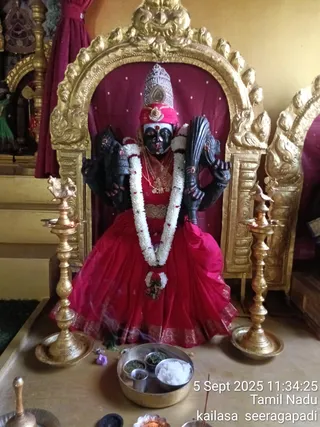A dark-complexioned deity murti of Kamakshi, seated in a gilded arched shrine with a deep red backdrop. The murti wears a silver crown, red and gold clothing with a red skirt bordered in gold, a white jasmine garland, and gold necklaces, and holds implements in multiple arms. Brass oil lamps flank the shrine, a brass thali with rice and green leaves sits in front, along with a small bell and incense smoke. A timestamp in the corner reads 5 Sept 2025 11:34:25, Tamil Nadu, kailasa seeragapadi.