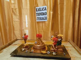 A photograph of a temple altar arrangement set against gold silk drapery. A white sign with bold black capitals reads KAILASA TRINIDAD & TOBAGO. On a dark stone slab sit three bronze-coloured murtis: a standing figure on the left, a tiered pedestal with a domed base at centre, and a seated deity on the right wearing a crown, holding a trident and adorned with red flower garlands and yellow blossoms. Scattered white and yellow flowers lie around the base, and the white marble floor is visible at the front.
