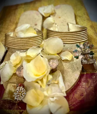 Close-up photograph of a pair of ceremonial gold sandals or padukas resting on a gold brocade cloth over a magenta and gold patterned fabric. Pale yellow rose petals are scattered over the footwear and cloth. Two gold-toned metal rings or bangle-like bases sit at the front of the sandal, and a small ornate jewelled object, possibly a miniature flower-shaped decoration with green and pink stones, rests at the right edge. No people are visible; the scene shows only the sacred footwear and floral offering.