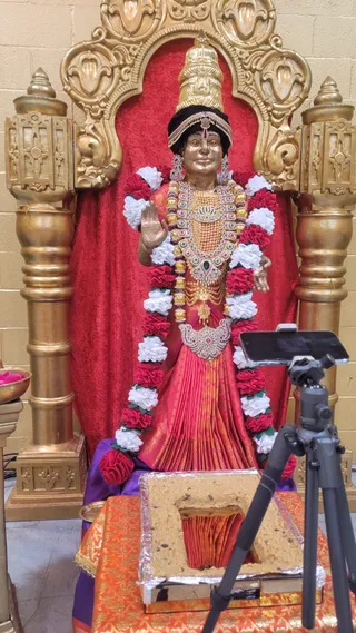 A photograph of a golden deity murti standing in an ornate gilded arch and carved gold pillars against a red velvet backdrop, in a plain cream-walled room. The figure wears a tall golden crown, a jewelled necklace and a pink-red silk sari, with a garland of red and white flowers draped from the shoulders. The murti's right hand is raised in abhaya mudra. In the foreground, a rectangular block of sweet offering, possibly prasadam, rests on an orange and gold brocade cloth beside a tripod-mounted phone. A small brass bowl of petals sits at the left edge.