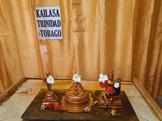 A photograph of a small altar display in front of gold draped fabric. A white paper sign with bold black capitals reads KAILASA TRINIDAD & TOBAGO. On a dark stone slab sit three gold-coloured metal figures: a small standing figure of a bald man in a dhoti on the left, a tiered pyramid-like sri yantra style base with a white plumeria flower on top in the centre, and a seated deity murti with a tall crown and a garland of red flowers on the right. White plumeria flowers and red ixora blossoms are scattered on the stone and placed on the figures. No people are visible.