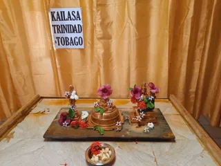A photograph of a ritual shrine set up on a wooden board before gold curtains. Three small golden metal figures stand or sit on the board: a standing figure on the left, a tiered golden peetha or pedestal form in the centre decorated with pink hibiscus-like flowers and white plumeria, and a seated deity murti on the right with a crown-like head ornament, garlanded with red flowers. Scattered red ixora and white blossoms surround them. A small steel bowl of sliced banana or similar offering sits on the floor in front. A paper sign reading KAILASA TRINIDAD & TOBAGO is taped to the curtain. No people are visible.
