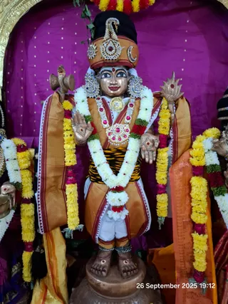 A standing deity murti, a gilded statue dressed in an orange and gold brocade garment with a striped tiger-pattern waistband, wears a tall ornate crown with a crescent moon and jewelled ornaments. It is garlanded with white jasmine, yellow marigold and red rose strands, and wears a large jewelled necklace and earrings. It stands before a magenta draped backdrop on a pedestal. No physical people are visible; the photo shows a statue with a timestamp reading 26 September 2025 19:12 in the corner.