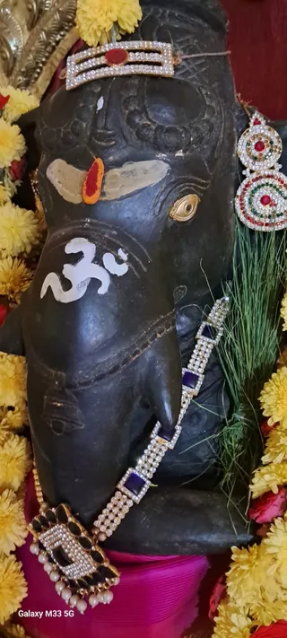 Close-up photograph of a dark black stone deity murti. The face has a white Om symbol painted on it, a red tilak with white markings on the forehead, and a gold-rimmed eye. The murti is adorned with a jewelled gold crown or headpiece with red and white stones, large jewelled earrings in red, green and white, a blue and white stone-studded necklace and a pendant with pearl drops. Yellow and orange marigold garlands surround it, along with green grass blades and a pink cloth below. No people are visible.