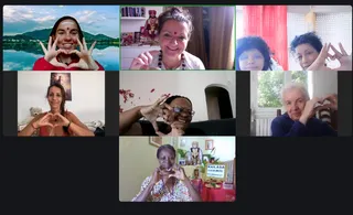 A seven-panel video call grid of participants making heart gestures with their hands. Top left, a smiling woman with a white forehead mark and red top stands before a lake and mountain backdrop. Top centre, a smiling woman with a red bindi and beaded necklace sits before a bookshelf holding a small figure of a turbaned, bearded figure in ornate red and gold clothing; this figure is a depiction on a shelf, not a clearly identifiable person. Top right, two women make hand gestures in front of red curtains. Middle row, a woman in an orange top sits on a bed with a teddy bear, a woman with glasses sits on a sofa against a wall, and an older woman with white hair in a navy top sits by a window. Bottom centre, a woman in a patterned top makes a heart gesture beside an altar with a framed deity image, flowers, a kalasha-style offering and a sign reading KAILASA GUADELOUPE.
