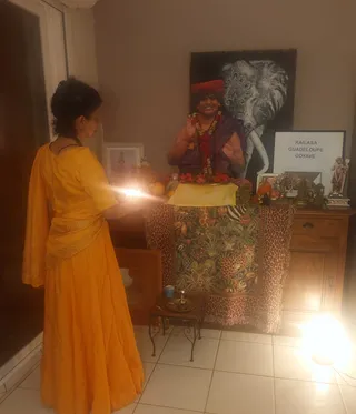 A woman in a flowing saffron-yellow dress and matching wrap stands at a home altar, holding a lit oil lamp toward a shrine. On the altar, draped with a leopard-and-jungle patterned cloth, a framed photograph shows a smiling figure with a red turban, red and orange garlands and a purple shawl, with a large elephant artwork behind. Nearby stand a brass plate with a small vessel, oranges, gold deity figurines and a framed sign reading KAILASA GUADELOUPE GOYAVE. A wooden cabinet sits to the right in a tiled room.