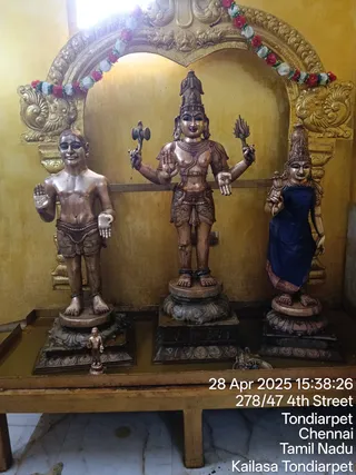 Three life-size wooden statues stand on a low wooden platform in front of a golden-yellow wall with an ornate gilded arch above. A garland of red and white flowers hangs along the arch. The central figure is a four-armed deity murti with a tall crown, holding a trident-like object in one upper hand and a lotus-like object in the other, with the lower hands in abhaya and varada poses. To its left stands a bare-chested male figure with a waist cloth and a raised hand. To its right stands a female figure in a dark blue garment with a crown and a lotus-like object. A small brass figure sits on the platform in front. The image is a photograph with a date and location stamp reading 28 Apr 2025 and Tondiarpet, Chennai.