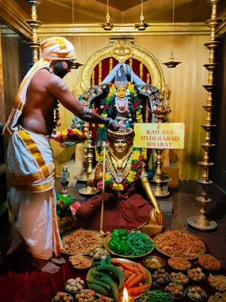 A photograph of a temple shrine where a priest in a white dhoti with gold border and a white-and-gold turban stands bare-chested with sacred ash on his body, offering a tray of flowers over a golden seated murti. The seated figure wears a crown, a red garment and many garlands. Behind it stands a dark deity murti in ornate silver-and-jewelled dress with a crown and marigold garlands, framed by a golden arch. Brass lamps stand on both sides, and a yellow sign reads KAILASA HYDERABAD BHARAT. In front, many plates hold vegetables, sweets, savouries and fried snacks, with an oil lamp burning at the bottom edge.