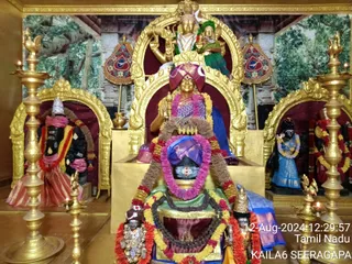 A photograph of a temple shrine interior with gold-framed arches and golden oil lamps on stands in the foreground. A large central deity murti sits on a gold throne beneath a gold arch, with a pink-and-purple garland and a purple headcloth, a red-and-purple garland draped across a tall Shiva lingam with three horizontal lines and a rudraksha bead garland around its base. Smaller black stone deity murtis flank the scene, one in a blue sari with a flower garland and another with a red cloth, all in front of painted forest backdrops. Small flames burn in the lamps. There are no physical people visible.
